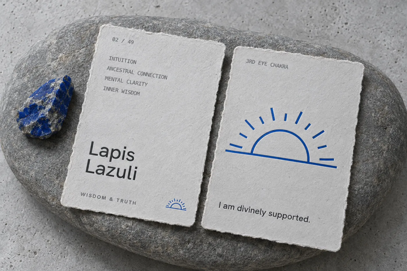 Concept photograph of two torn-edge paper stone cards on a grey river stone beside a raw lapis lazuli: the front reads 02 of 49, Lapis Lazuli, Wisdom and Truth, and the back carries a large blue sunrise and the line I am divinely supported