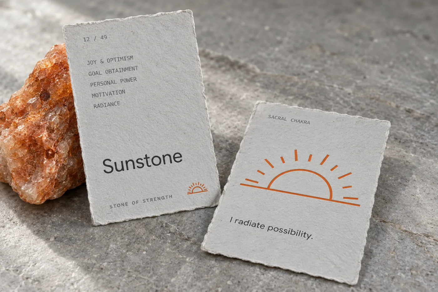 Concept photograph of two torn-edge paper stone cards beside raw sunstone: the front reads 12 of 49, Sunstone, Stone of Strength, and the back carries an orange sunrise and the line I radiate possibility