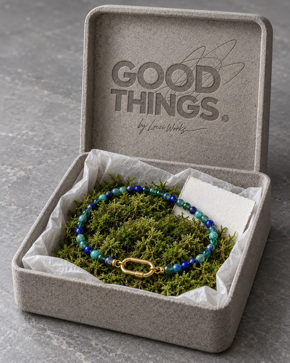 Concept render of an open molded mushroom box with the GOOD THINGS mark embossed in the lid, rice paper inside and a lapis, turquoise and green strand with a gold clasp resting on a bed of moss