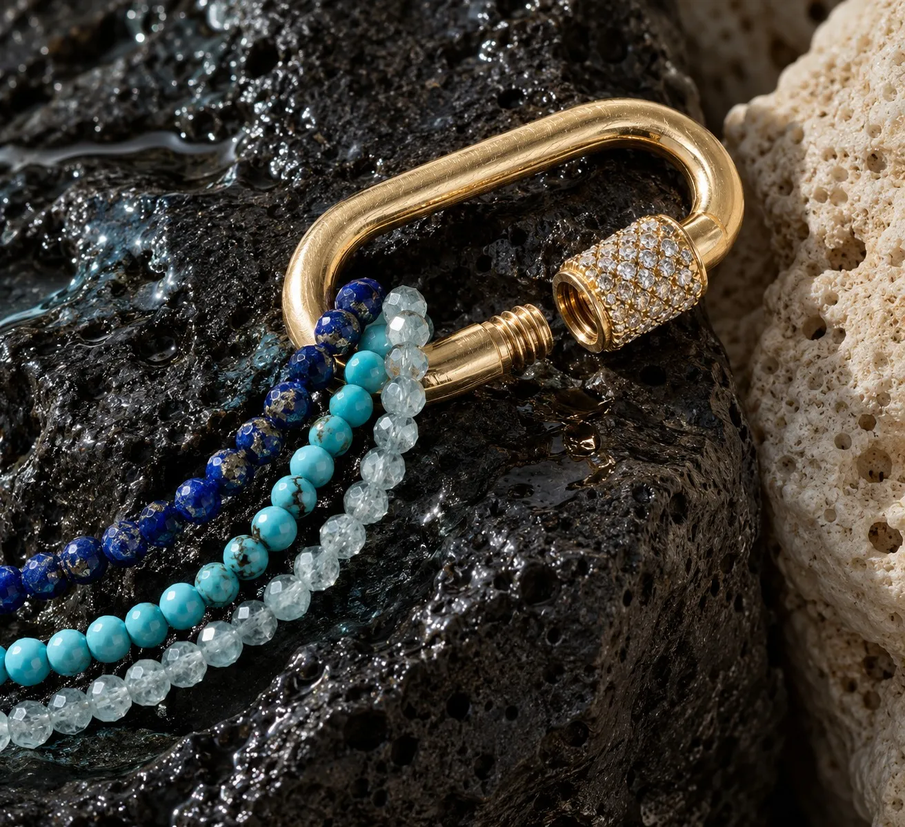 Gold pavé carabiner holding lapis and turquoise strands on dark volcanic stone